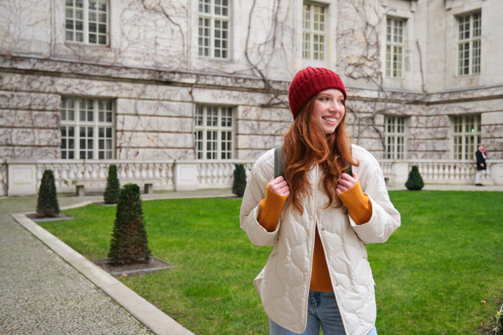 Young smiling redhead woman walking beautiful city attractions wearing backpack red hat coat